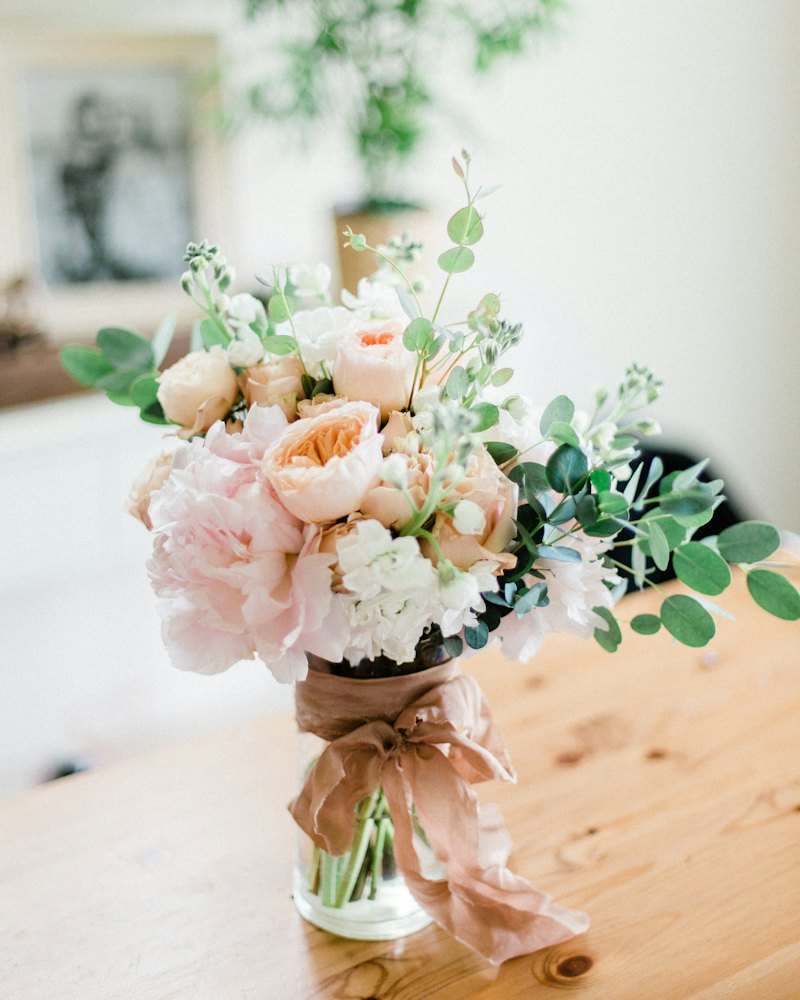 Bouquet aux tons poudrés, roses pâles et feuillage, dans un vase sur une table en bois